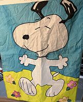 Photo of a vintage Snoopy garden flag depicting Snoopy dancing on a blue sky and green grass background with flowers.