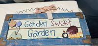 Front view of a rectangular wooden garden hanging plaque with a blue distressed frame, wire hanger, painted text 'Garden Sweet Garden' and decorative tulips and ladybug with fabric bow.