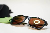 Front angle of Bolle 527 sunglasses showing matte black frame and brown emerald polarized lenses with green reflective coating, placed next to green cleaning cloth and black case in background.