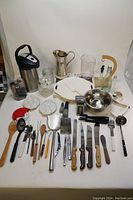 Full view of all kitchen items arranged on table, showing coffee thermos, pitchers, cookie jar, oil bottles, knives, and utensils