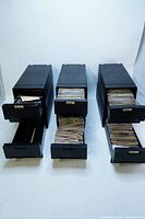 Photo showing six black drawers partially open revealing a large quantity of stacked papers inside, identified as original music album prints and liner notes.