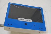 Front view of the American Dynamics 7-inch MiniMonitor showing blue bezel, built-in camera lens, and screen with protective film.