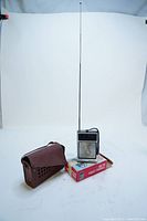 Photo of the handheld radio standing next to its leather carrying case and original packaging box. The radio is upright with antenna fully extended.