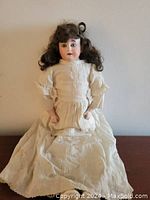 Full front view of the antique bisque doll seated, showing original white dress and brown hair.