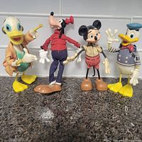 Four Disney figures including Scrooge McDuck, Goofy, Mickey Mouse, and Donald Duck lined up on countertop, showing full body details and wear.