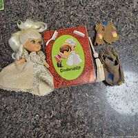 Wide view of all contents of the lot on countertop: Cinderella doll dressed in cream and silver gown, Cinderiddle storybook, brown vest, broom, and bundle of straw.