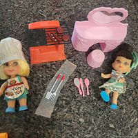 Photo of Sizzly Friddle Yum Yum doll in chef outfit, plastic barbecue grill with two patties, grilling tools, Pretty Priddle doll, and pink dressing table with accessories.