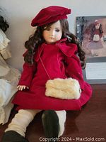 Full frontal view of Eaton Beauty Doll seated, wearing red velvet coat dress, beret with white pom-pom, faux fur muff, cream leg coverings, and black shoes.