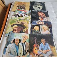Full view of eight LP record covers laid out on a bed, showing artists' names and album titles including John Denver, Kenny Rogers, Dolly Parton, and others.