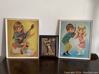 Three framed artworks on a wooden surface: two 'Big Eyed Kids' paintings and one 'Pity Kitty at the Dump' cat painting.