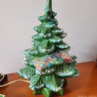 Green gloss-finished ceramic Christmas tree with white snow effect, standing on a wooden surface, with sealed multi-colored plastic lights attached.