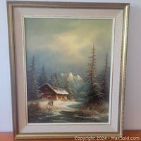 Full view of the framed oil painting showing the winter cabin scene with snow and pine trees under a cloudy sky.