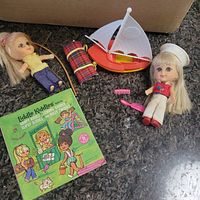 Photo showing two Liddle Kiddles dolls, sailboat toy, rolled-up plaid item, pink comb, and green booklet on dark surface