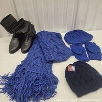 Black boots, blue scarf, two blue knitted hats, blue fingerless gloves, black knitted hat with tag shown together on white background.