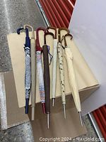 Seven folded umbrellas lying on a beige surface, showing variety in colors and handles arranged side-by-side.