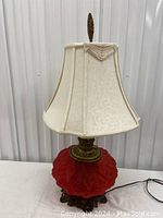 Full view of the lamp with red embossed glass base and white fabric shade on a white table with metal base visible.
