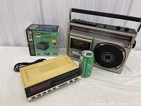 Photo showing all three items: vintage cassette radio, digital alarm clock, and boxed transformer alongside a soda can for scale.