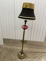 Full view of the vintage floor lamp showing brass base, cranberry glass accent, and black fabric shade with gold fringe trim.