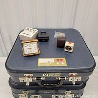 Two identical blue vintage suitcases stacked, with travel clocks and small accessories on top including a gold-tone clock, black travel clock in a clear cube, small black rectangular item, and packaged small parts.