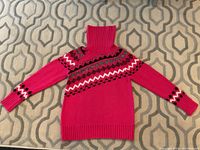 Front view of bright pink turtleneck sweater with multi-color geometric pattern on chest and sleeves, laid flat on patterned surface.