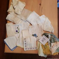Top view of 24 vintage handkerchiefs spread on table showing various embroidered floral designs, lace and crochet trims, in assorted white, cream, and pastel tones. Includes visible tags for lot identification.