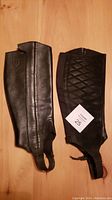 Pair of black Ariat leather half chaps laid flat on floor showing front and rear sides.