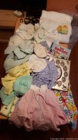 Photo showing vintage knitted baby clothes, bonnets, sweaters, small baby shoes, a tiny moccasin, baby blankets including a handwoven one, children's book, picture frame, and fabric dresses in pastel colors