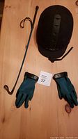 Photo of black riding crop, pair of teal riding gloves and black equestrian helmet on wooden surface.