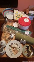 Assorted kitchen and household items on table including plates, placemats, salad spinner, glassware, decorative plates with rooster and cheese motifs, totes, basket, tray, and pet dish.