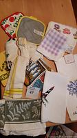 Top view of assorted kitchen linens featuring printed tea towels, potholders, and terry towels arranged on a wooden table.