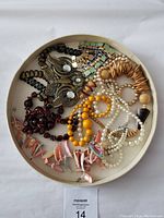 Image showing various necklaces laid inside a circular tray, showcasing different colors, styles, and materials including beads, shell pieces, and metallic elements.