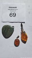 Three pendants on a flat surface with auction tag 69 visible: green labradorite triangular pendant, angular amber teardrop pendant, oval amber pendant