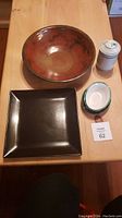 Photo showing four items: large brown shiny serving bowl, large satin black square platter, white cylindrical lidded porcelain jar, and colorful handpainted Mexican soap dish.