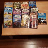13 volumes of the Redwall children's fantasy book series by Brian Jacques arranged on a wooden surface, showing front covers with colorful medieval animal protagonist illustrations.