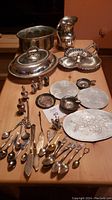 Overview of all silver plated spoons, water jug, salt and pepper shakers, serving dishes, aluminum coasters, brass and silvertone planter and French wine cup arranged on a table.