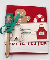 Front view of the gift set showing red apron with gingerbread man and candy cane design, wooden spatula with gingerbread man silicone head, and metal gingerbread man cookie cutter all wrapped in original packaging band.
