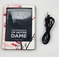 Front of KOBO N905B Touch Edition eReader showing 'The Hunchback of Notre Dame' eBook cover on screen and included USB charging cable.