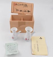 Two white ceramic soy sauce jars with light pink floral patterns in front of open wooden box with Japanese calligraphy on lid and two certificate papers.