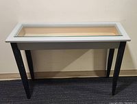 Front view of sofa table showing black legs and rectangular frame with glass top revealing storage compartment underneath