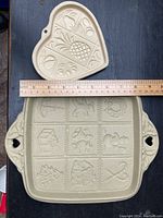 Photo showing two ceramic cookie molds: one heart-shaped, one square with Christmas designs. A wooden ruler is placed on top for size reference.