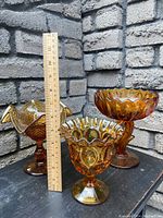 Three amber glass decorative bowls of varying sizes, each with a footed pedestal base and ruffled edges, positioned beside a 12-inch ruler for size reference against a grey brick wall background.