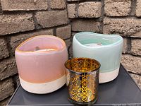Photo of two ceramic-scented candles in pink and green containers along with a decorative gold metallic votive glass showing ornate cutout design.