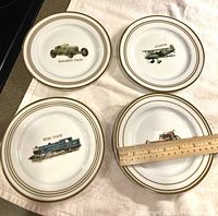 Four white cocktail plates shown from the front, each with a different vintage vehicle illustration and gold rim. A wooden ruler is placed on one plate indicating size.