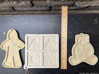 Photo showing two shaped cookie molds (gingerbread man and teddy bear) next to a square cookie mold with four holiday scenes, all beige colored and about 7 inches tall, with a wooden ruler for scale.