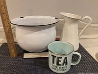 Photo shows a white enamel wash basin with a black rim, a teal enamel tea mug with black text, and a white ceramic jug with handle marked IKEA.