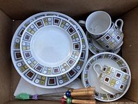 Top down view inside box showing stacked dinner plates, two cups and saucers, and skewers with wooden handles