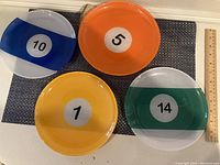 Four round glass plates arranged on a dark mat, each designed as a numbered pool ball: yellow 1, orange 5, blue 10, and green 14. A wooden ruler indicates plate diameter around 6-7 inches.