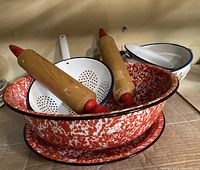Photo showing large red splatter enamel bowl, white enamel colander with a blue rim, two vintage wooden rolling pins with red handles, and parts of the white enamel pots.
