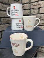 Photo showing four Tim Horton coffee mugs: two white mugs with red-black text and two cream-colored mugs with classic logo and coffee cup designs.