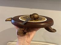 Side view showing the wooden turtle-shaped cheese board with detailed gold metal turtle limbs and ceramic insert. The turtle-shaped spreader is resting on top with handle in the tail area.